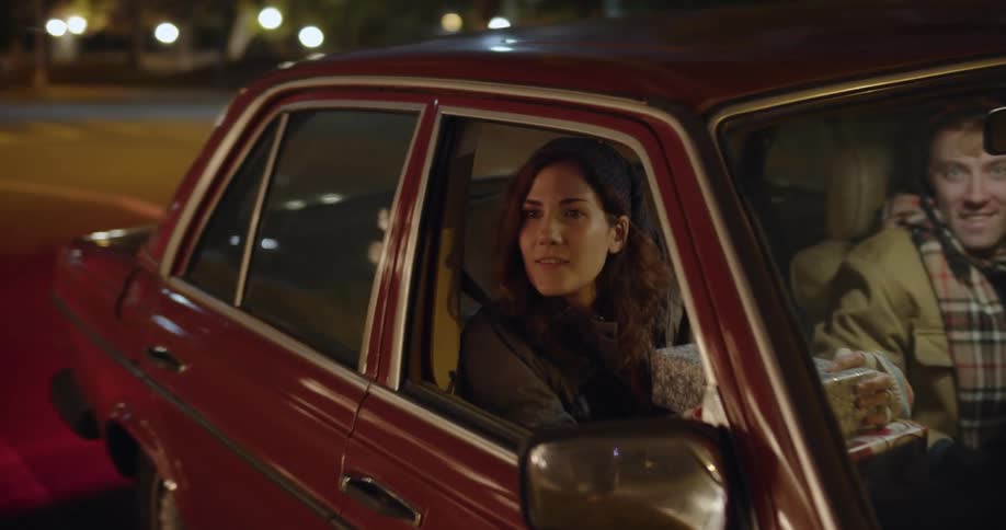 A woman is sitting in the passenger seat of an automobile with a smile on her face and a wrapped present in her lap as a man is driving and they slow down on the street