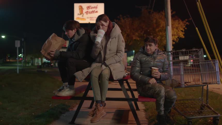 Three teenagers two boys and a girl sitting on a picnic table in a park and waiting for a bus while chatting and enjoying fast food and some drinks