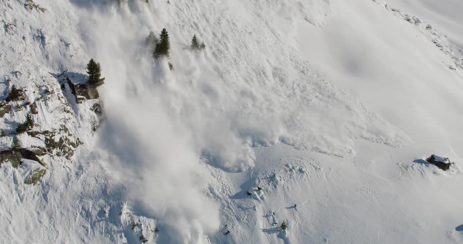 Aerial footage of a snow avalanche moving down a mountain valley at high speeds engulfing trees rocks in its path