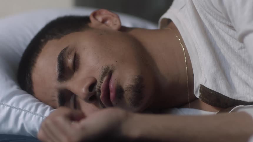 Teenage hispanic boy with minimum facial hair lying on bed with head on pillow slowly opens eyes awake