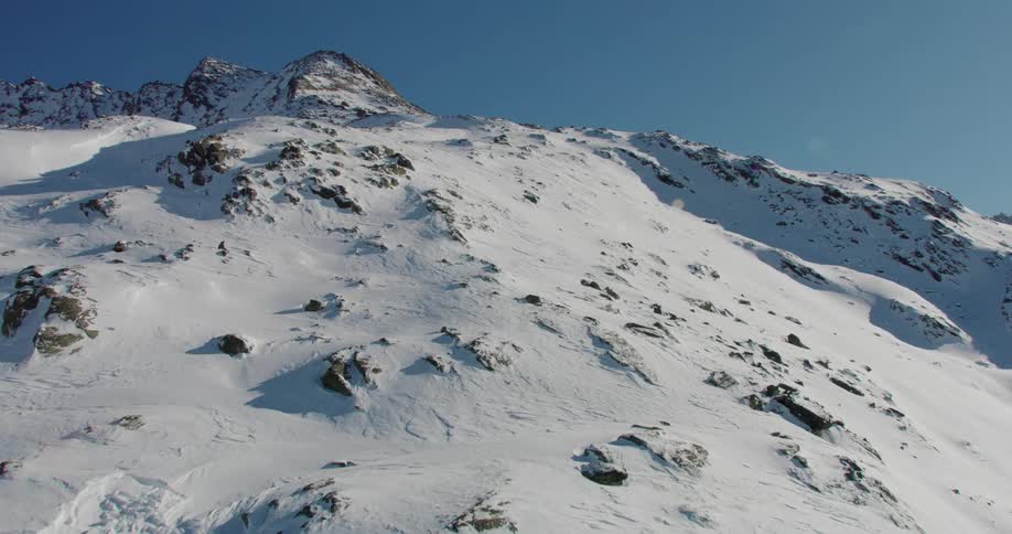 Wide aerial shot going up side of mountain peak covered in snow in midday with the sun shining