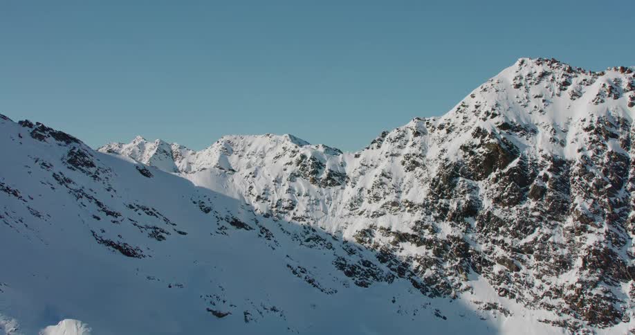 The sun shines on a snow covered mountain ridge and there are mountains in the distance under a clear blue sky
