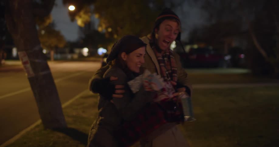 A man and a woman are walking on a neighborhood street and she is carrying wrapped presents as he puts his arm around her and spins her in a circle