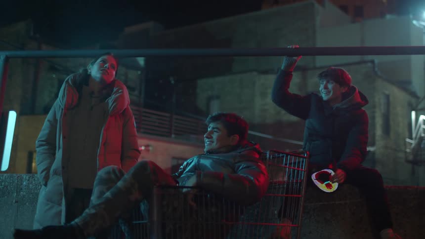 Friends talk outdoors at night near building with one holding a digital device and one sitting in grocery cart