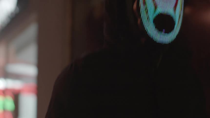 A man walks down a city street at night wearing a computer animated mask