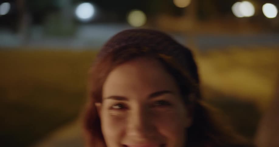 Woman smiles brightly as she walks toward person who hugs her in area brightly decorated by outdoor lights