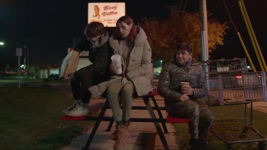 Friends sit on picnic table near street sharing fast food at night