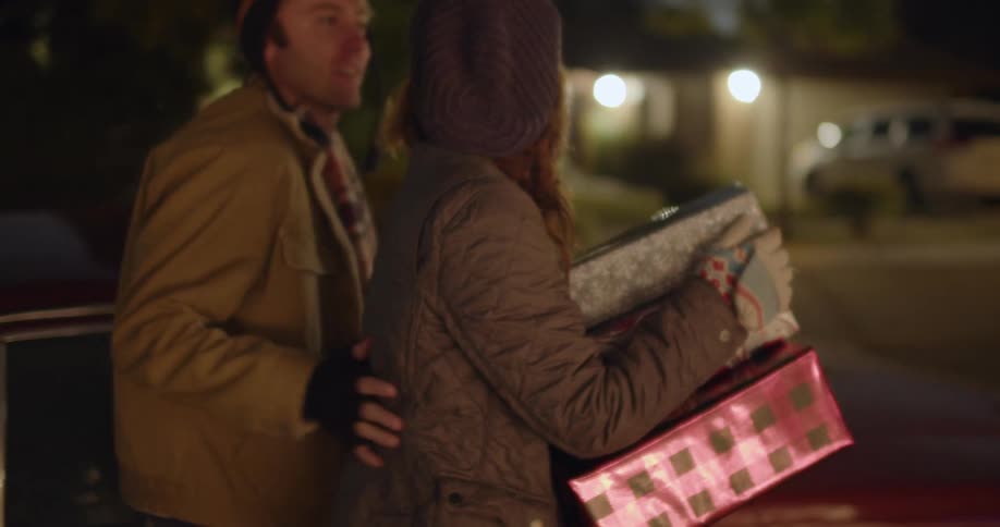 A woman gets out of the passenger side of an automobile carrying wrapped presents as a man kisses her and closes the car door as they walk away