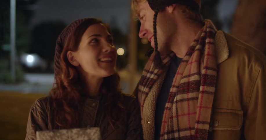 A man and a woman are standing on a neighborhood street and she is carrying wrapped presents as he has his arm around her and they kiss