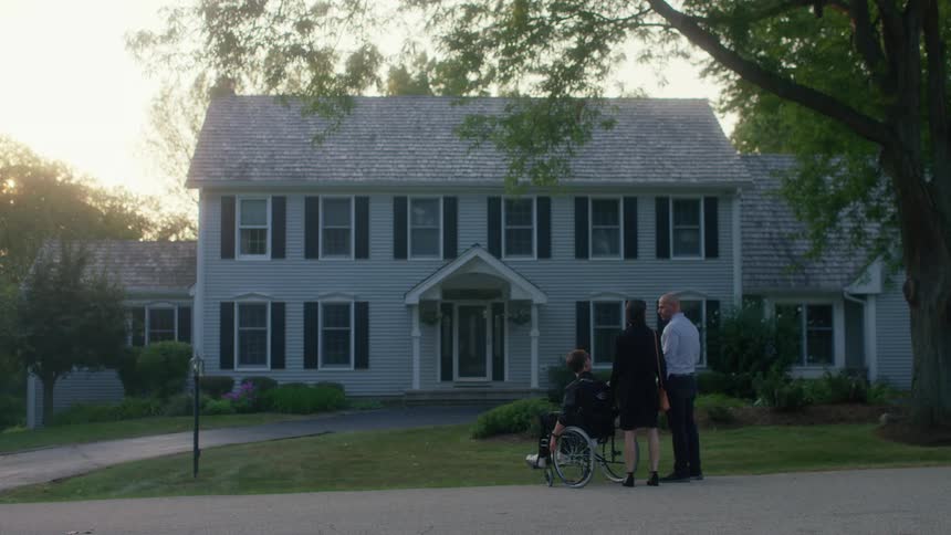 A big white house sits on a neighborhood street as a teenage boy in a wheelchair passes by on the sidewalk with his parents walking behind him