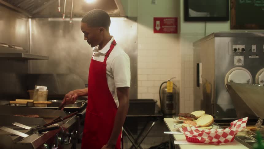 Young man cooking burgers on the restaurant girdle gets a light slap on the head by his coworker as a joke