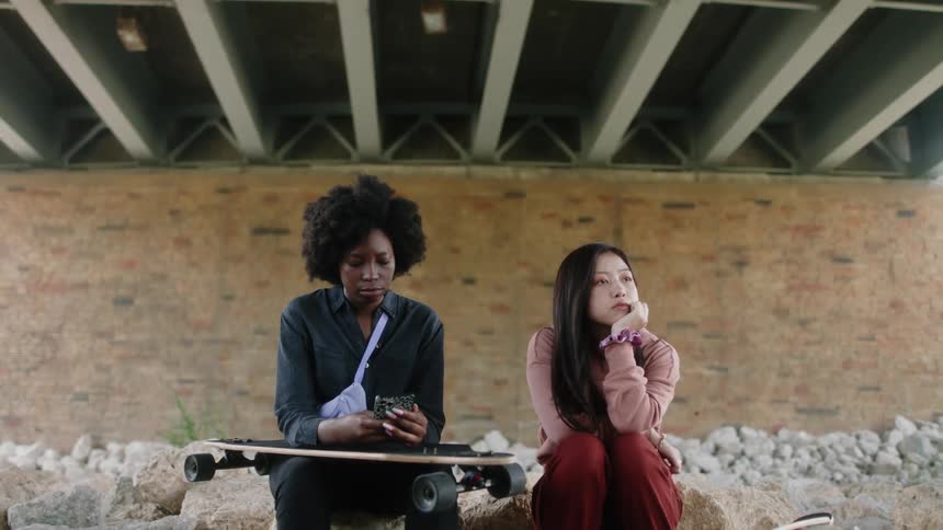 Two women sit under overpass as one holds a skateboard while talking and using a smartphone