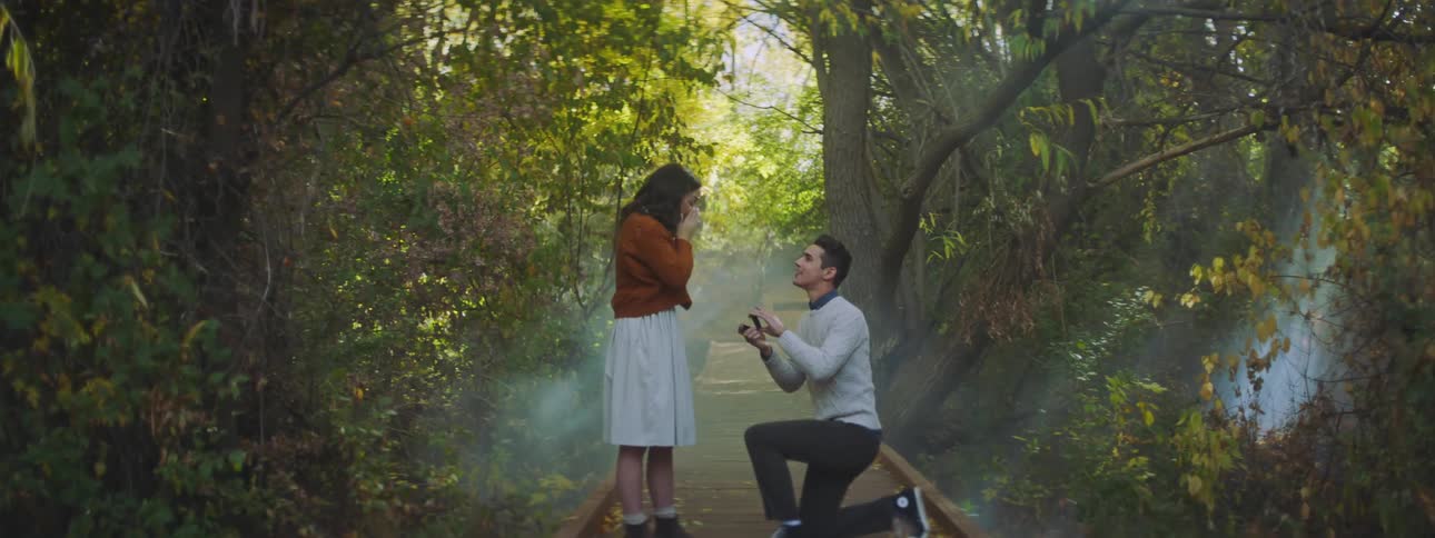 A man kneels and proposes to a woman on a pathway in a woodland area during the day