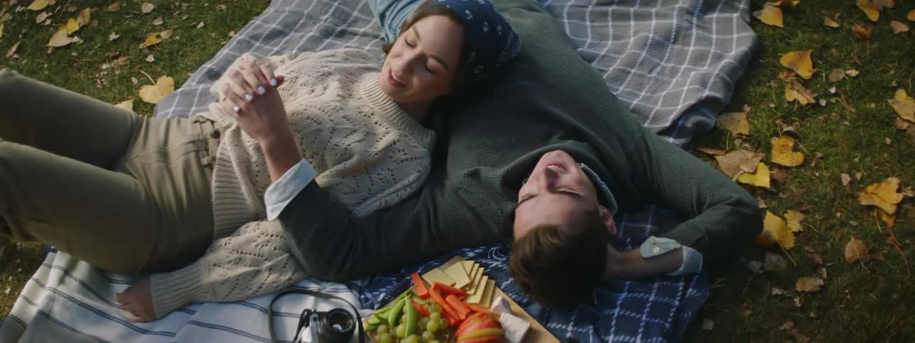 Couple lies together on picnic blanket near food on wooden chopping board