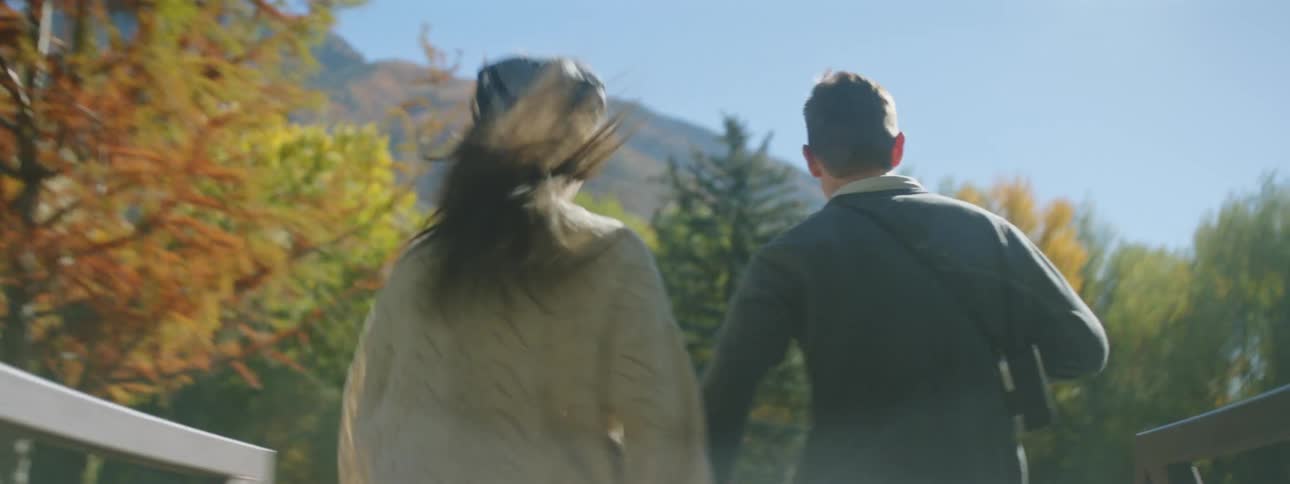 Couple runs hand in hand across bridge to field near woods at base of mountain