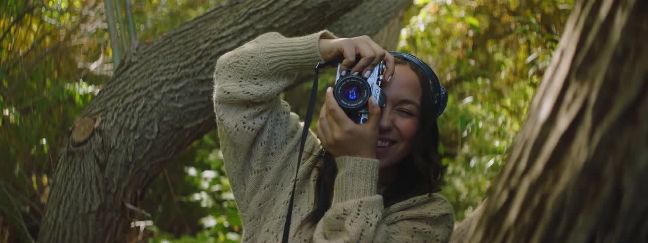 A woman is standing in the woods near trees smiling as she holds up her camera to take a picture