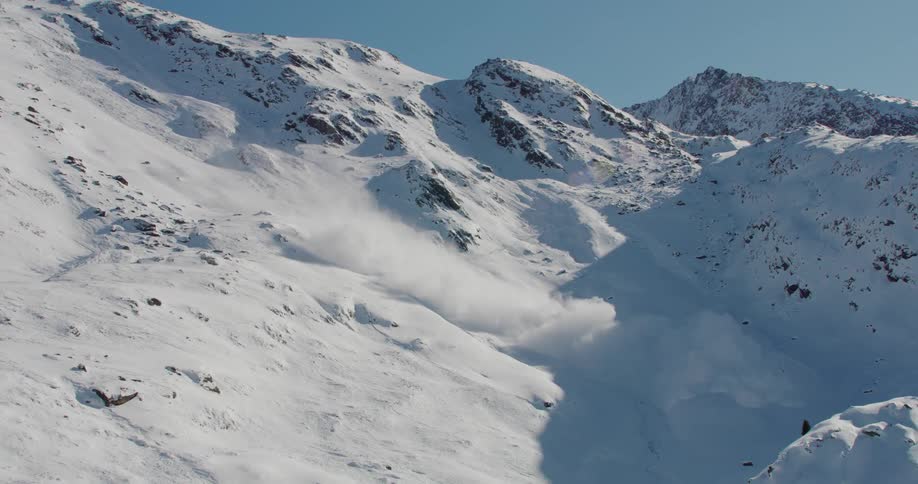 Panoramic view of snow rolling down the slope into the valley below during an avalanche in the mountains on a sunny day with clear blue skies