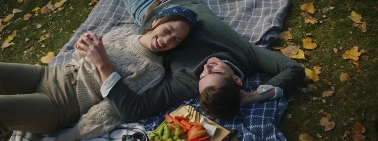 Couple lies on picnic blanket with food cuddling and talking
