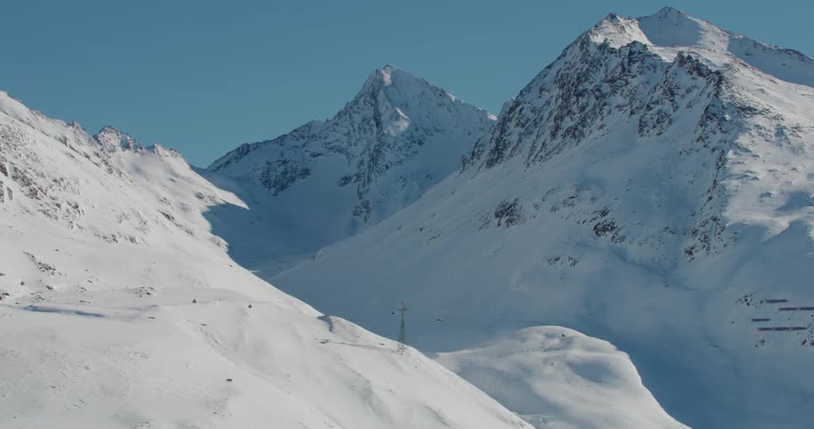 Ski lifts stand along the snow covered slope of a mountain range with peaks high in a clear blue sky