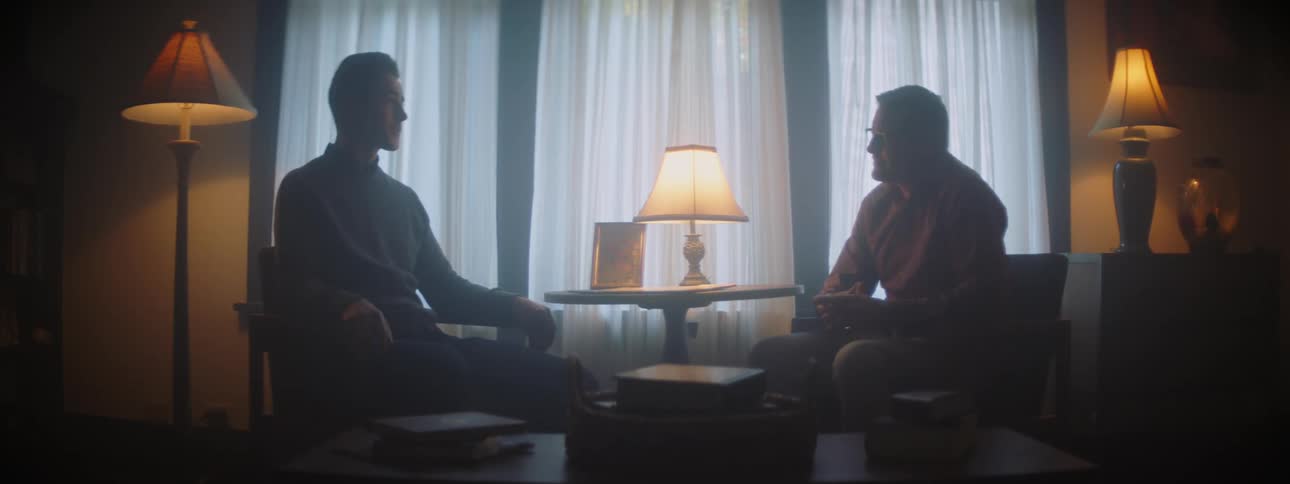 Men sitting in dim room finish discussion with a handshake and a hug