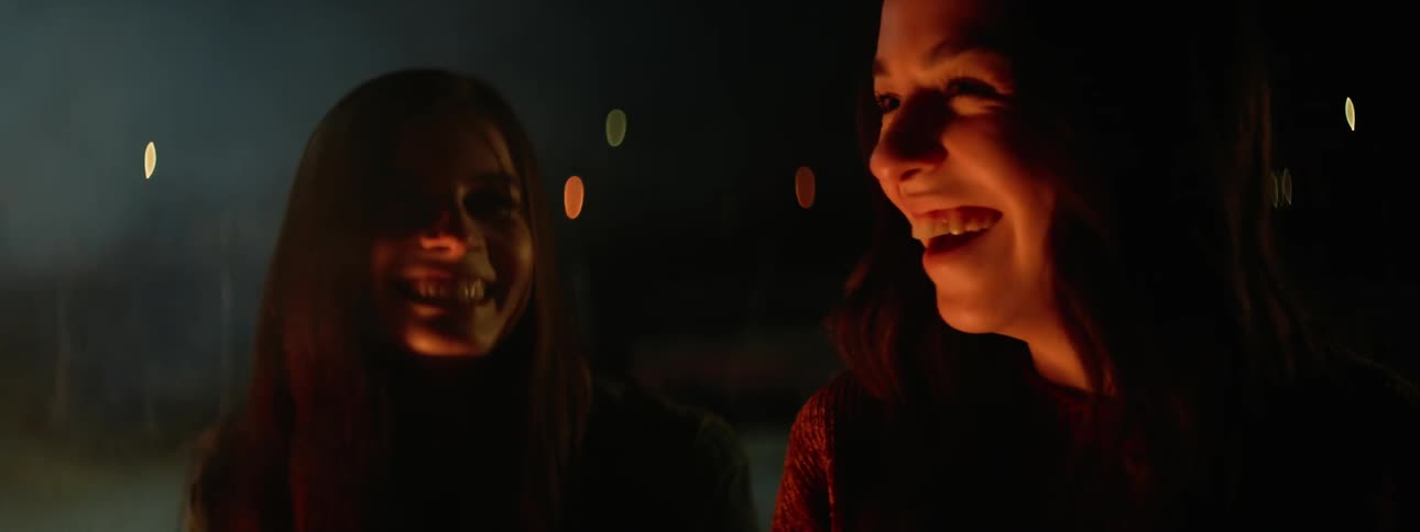Light shines on the faces of two women sitting outside at night laughing near a bonfire