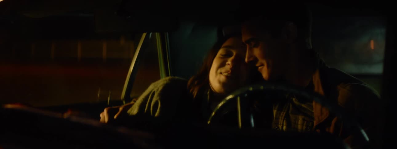 A man and a woman are sitting together in an automobile with his arm around her and their faces close together as they smile