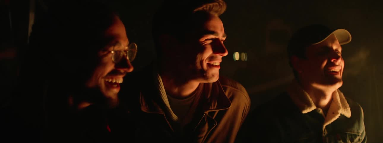 Three man laughing and smiling around a fire that reflects off their faces