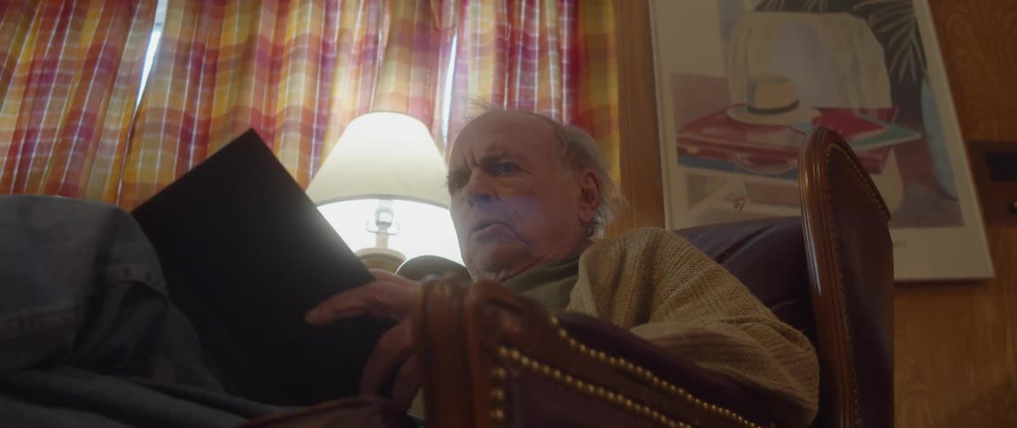 Elderly man with cardigan and pants looking scared sits in armchair reading a book in living room