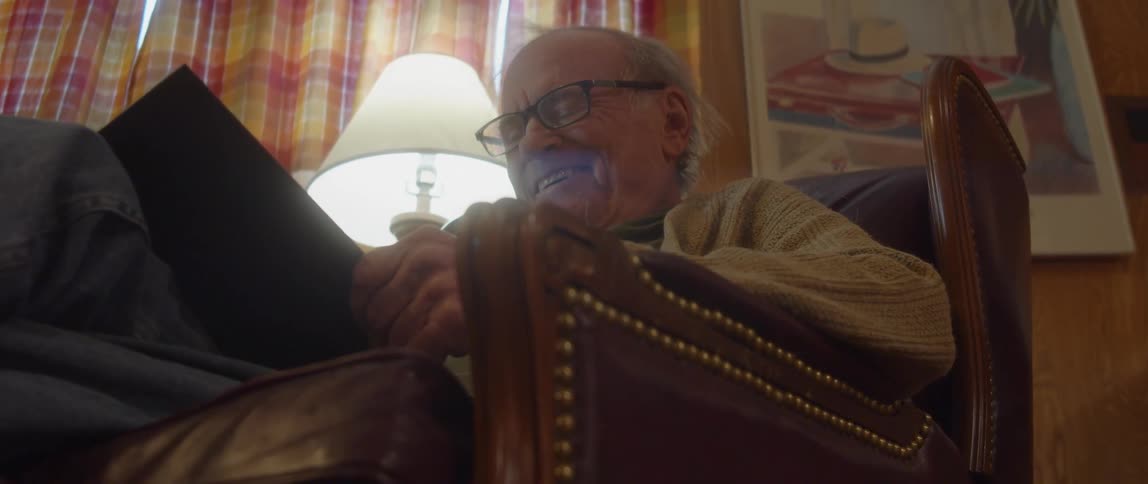 An elderly man is sitting in a chair in his living room holding a book in his hands and laughing