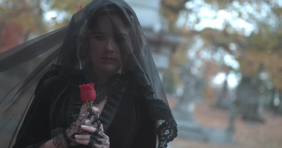 A woman dressed in a black dress and veil holds a red rose