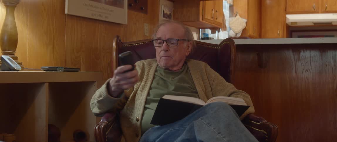 An elderly man is sitting in a chair in his living room reading a book and he reaches over to the table and answers his phone