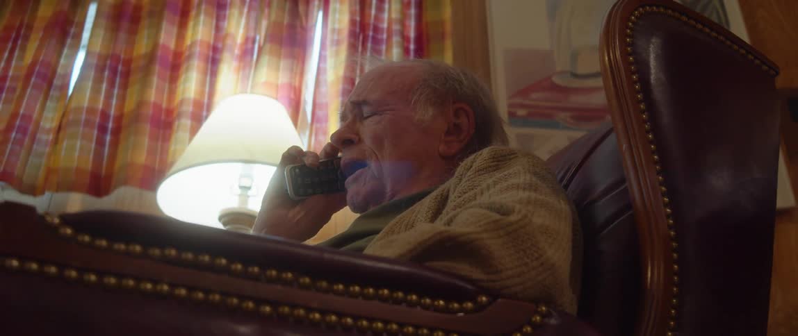 An elderly man is sitting in a chair in his living room with an open book in his lap smiling and laughing as he talks on the phone