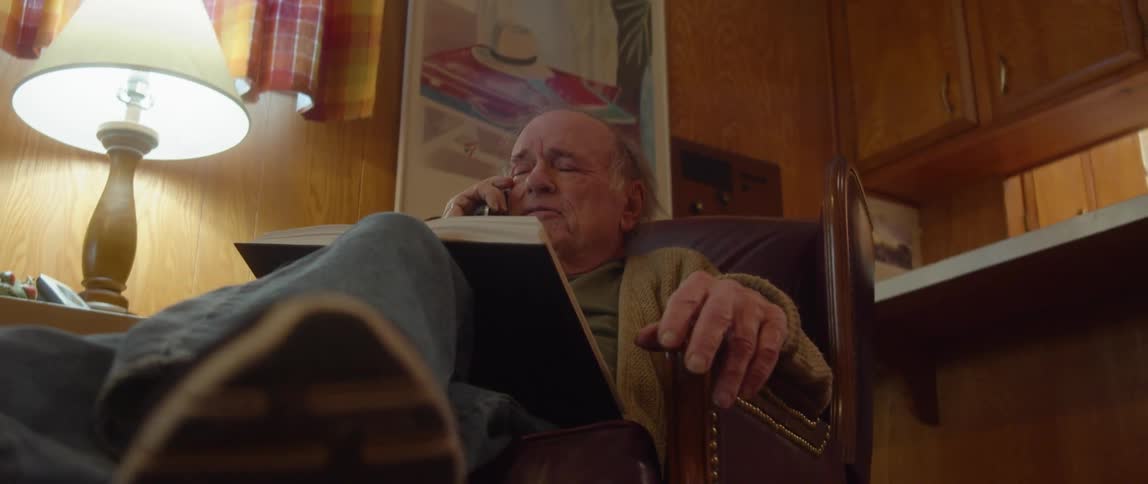 An elderly man is sitting in a chair in his living room with an open book in his lap as he talks on the phone