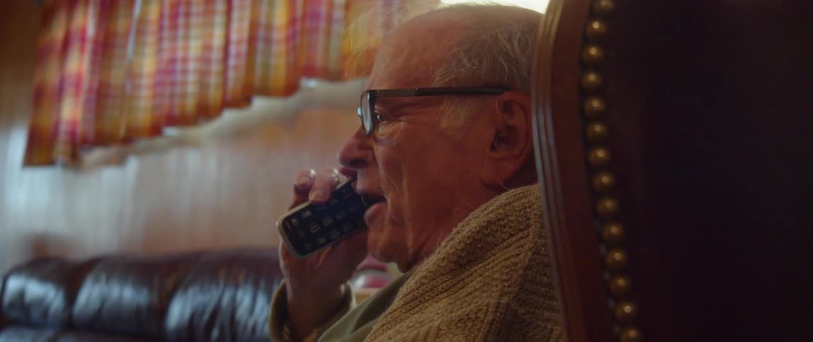 An elderly man is sitting in a chair in his living room smiling as he talks on the phone