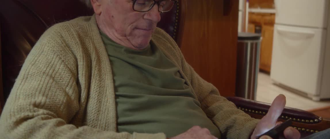 An elderly man is sitting in a chair in his living room holding his phone in his hand and scrolling with his finger