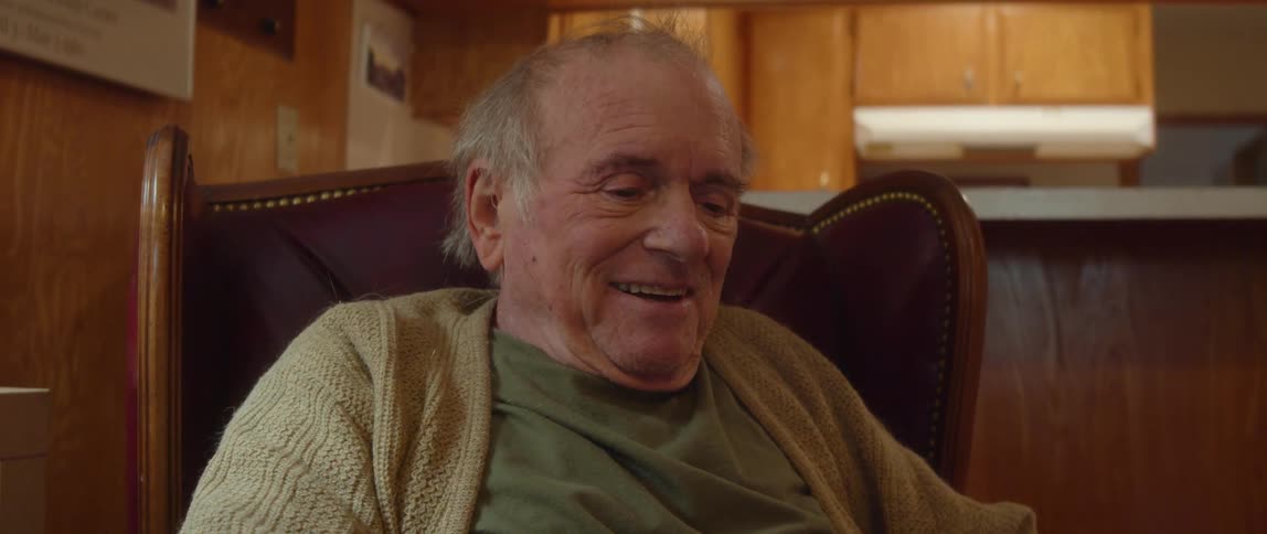 Elderly man with cardigan and green shirt sits in brown armchair talking and laughing