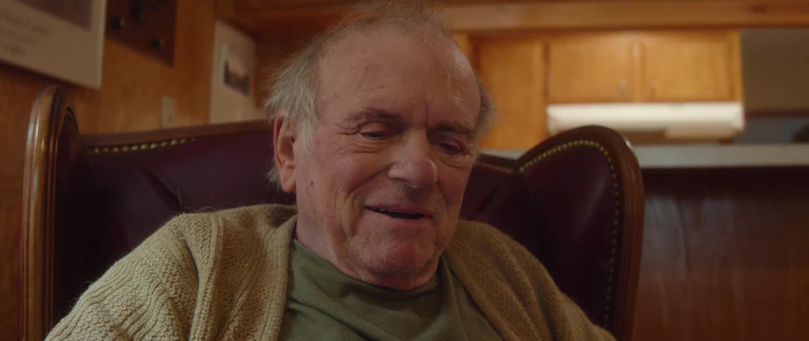 Elderly man with jeans green shirt and brown cardigan sitting in brown armchair talks and laughs in room