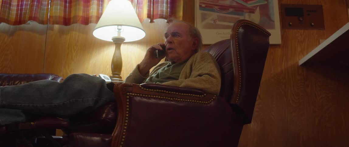 An elderly man is sitting in a chair in his living room as he reaches over to the table and answers his phone and takes off his glasses as he talks