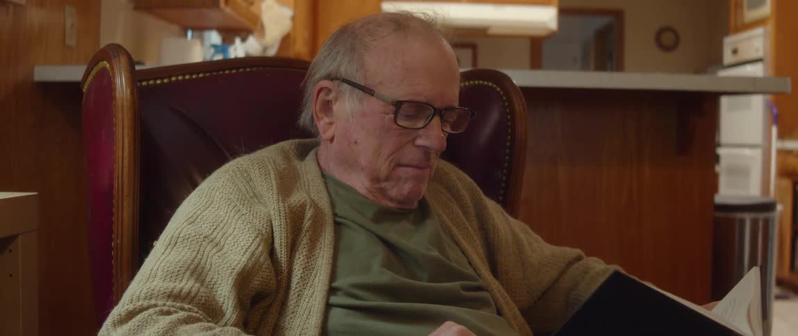 Elderly man with cardigan and shirt sits in brown armchair reading a book in room