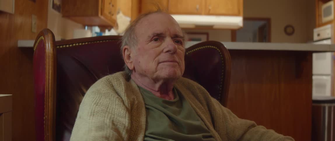 Elderly man sitting and reading in chair near kitchen takes off his eyeglasses