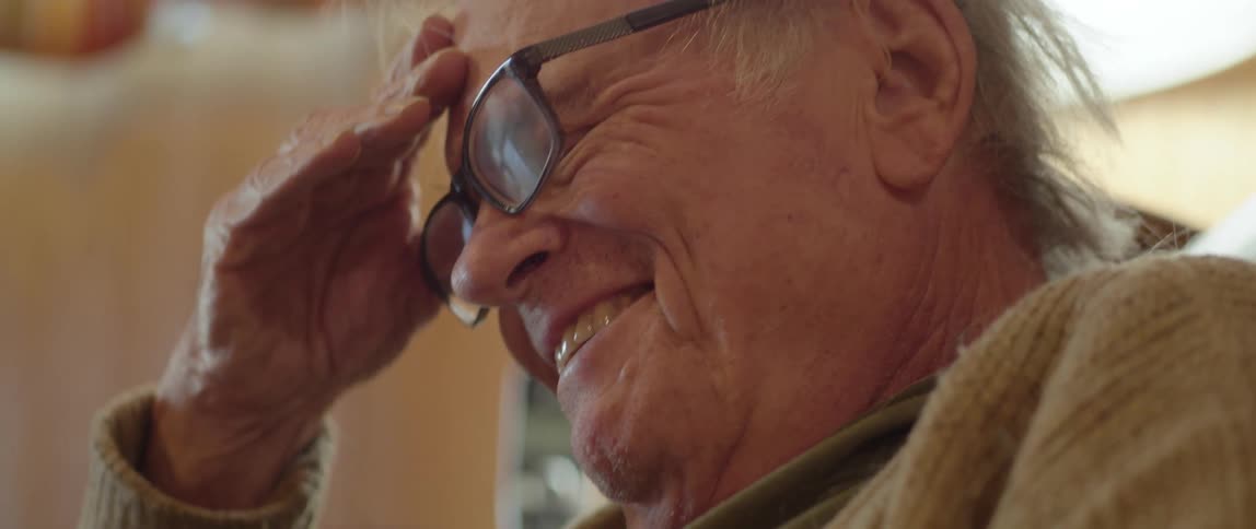 Elderly man with jeans green shirt and brown cardigan reads a book and laughs