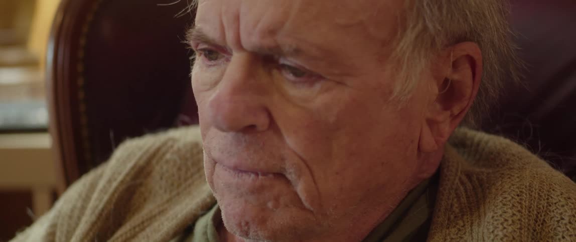 An elderly man is sitting in a chair in his living room and he takes off his glasses and pauses to think