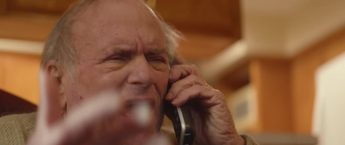 An elderly man is sitting in a chair talking on the phone and he appears shocked and angry as he moves his hand as he speaks