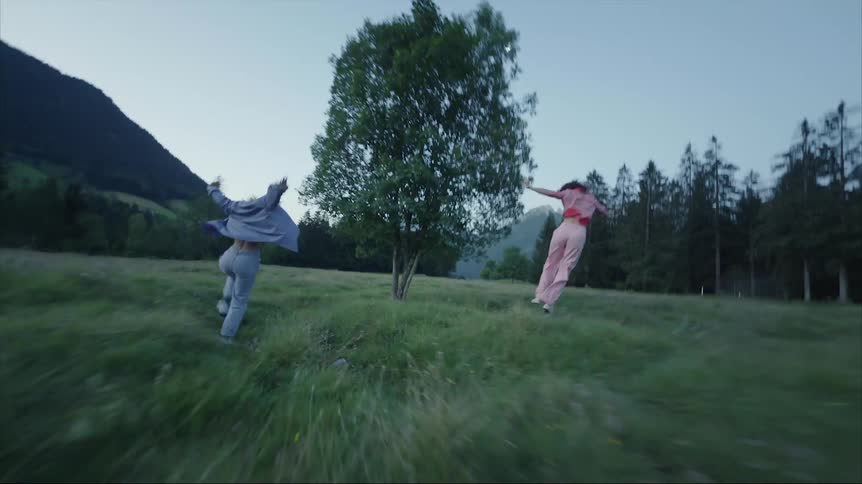 Two females one in pink the other in blue running in grassy clearing in a wooded mountain area