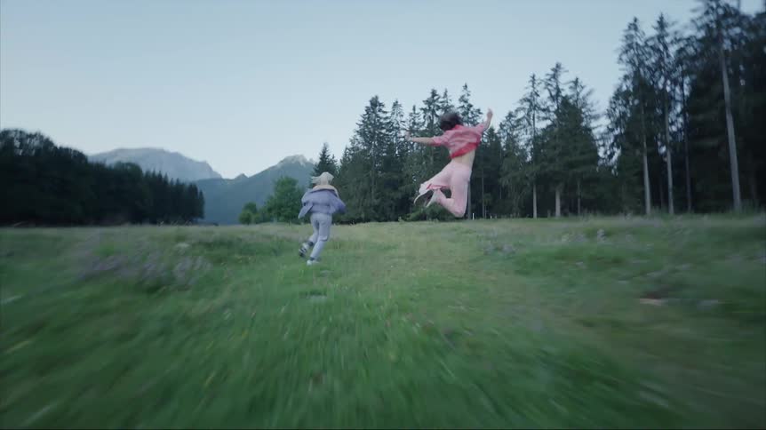 Two females run across field near woods with mountains in the distance