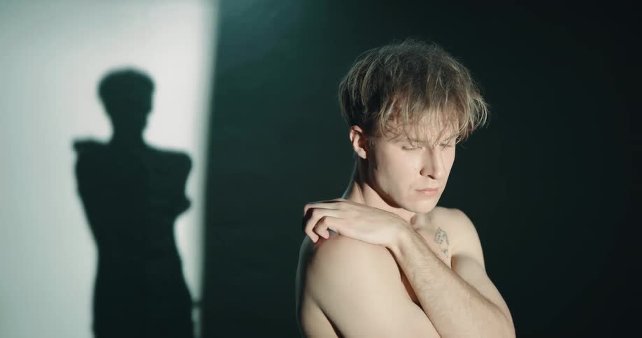 Shadow and light move across shirtless white emo man with his left hand touching his right shoulder looking vulnerable