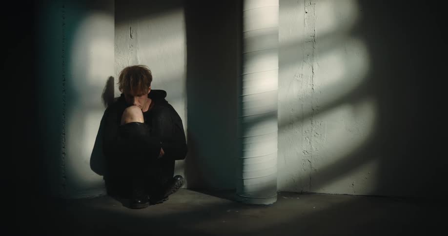 Young blonde androgynous man wearing all black crouching on floor in corner of warehouse sitting in light and surrounded by shadow