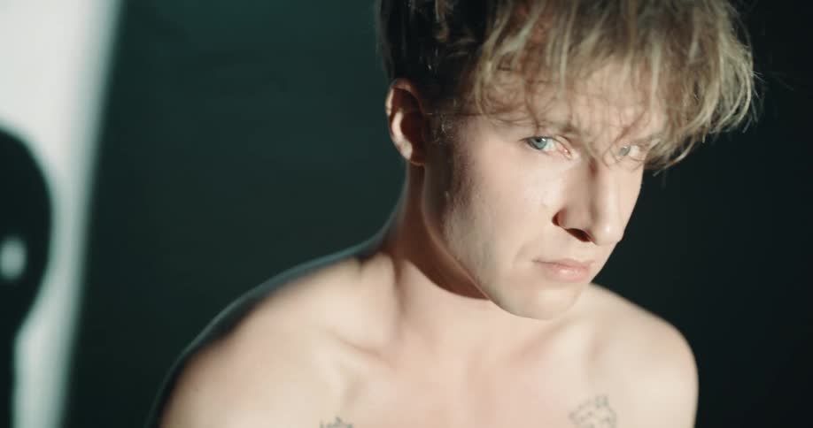 Topless shy man with tattoo stands and looks at camera in dark room