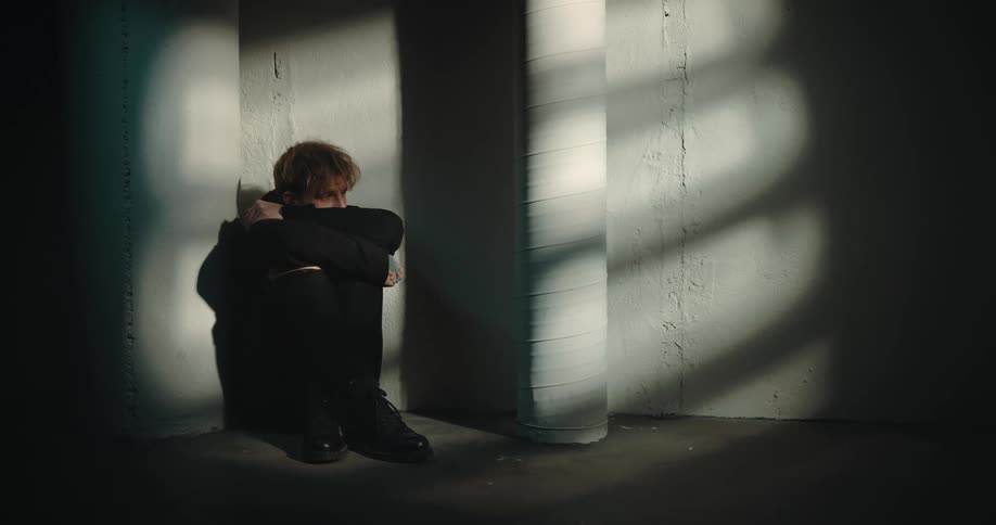 A man sits on the floor in an empty building with his knees pulled to his chest