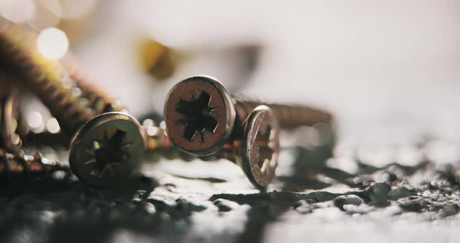 Extreme closeup of a pile of brass phillips head screws sitting on asphalt with shadows and out of focus fg elements moving across in slow motion